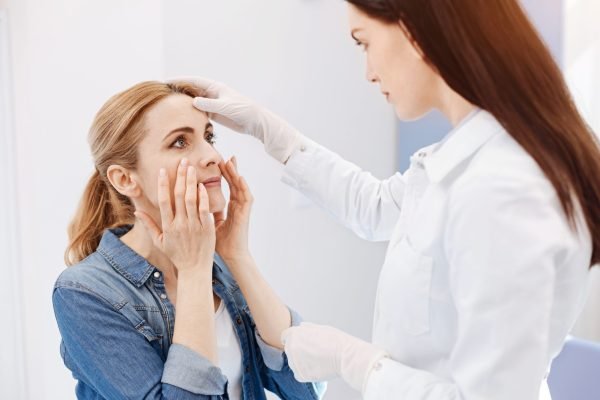 Look here. Beautiful good looking nice woman holding her cheeks and looking at the doctor while wanting to do a plastic surgery